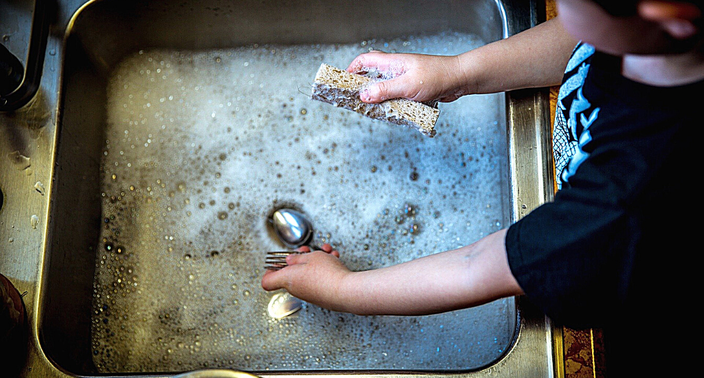 A child washes a fork and spoon in sudsy water in a metal sink.