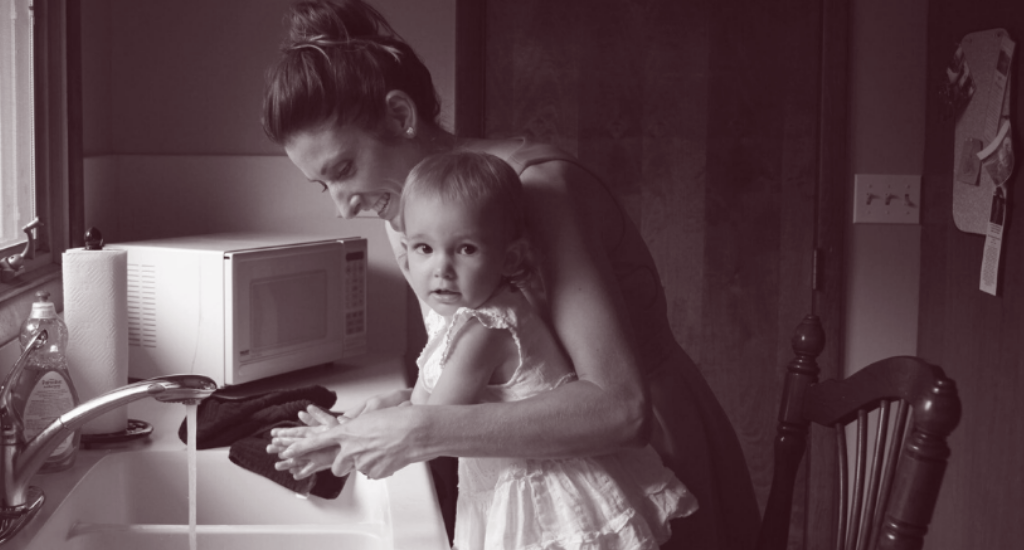 A young woman helps a toddler wash their hands at a kitchen sink.