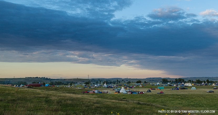A wide blue sky over a green field; in the distance sits a cluster of tents that make up the protest camp near the Dakota Access Pipeline.