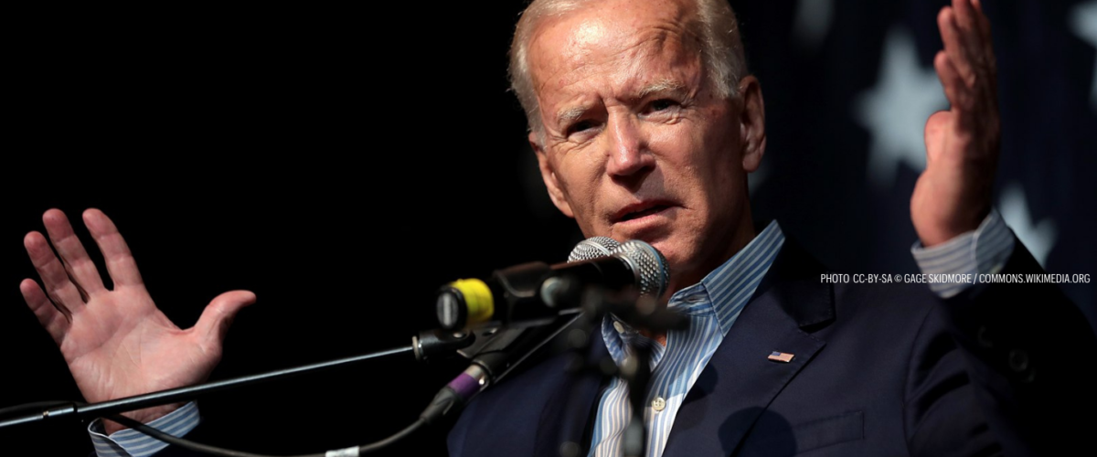 President Biden stands before a microphone with his arms extended.