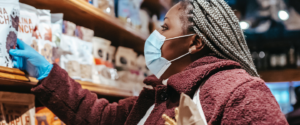 A Black woman wearing a surgical mask reaches for a product on a grocery store shelf.