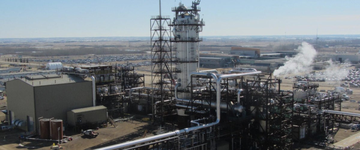 A carbon capture facility sits on a gray landscape. It has a gray main tower and it is covered in crawling black and silver pipes.