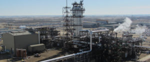 A carbon capture facility sits on a gray landscape. It has a gray main tower and it is covered in crawling black and silver pipes.