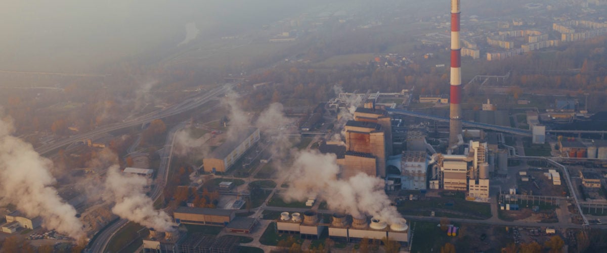 In an aerial view, smoke plumes and smoke stacks rise over a hazy urban landscape.