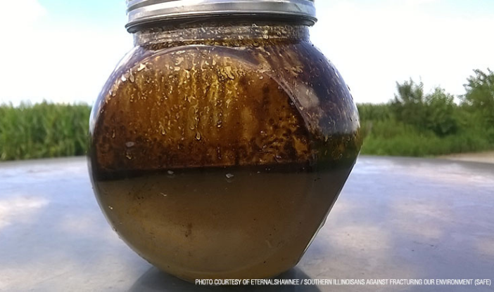 A jar of fracking sludge sits in a pool of murky water surrounded by greenery.