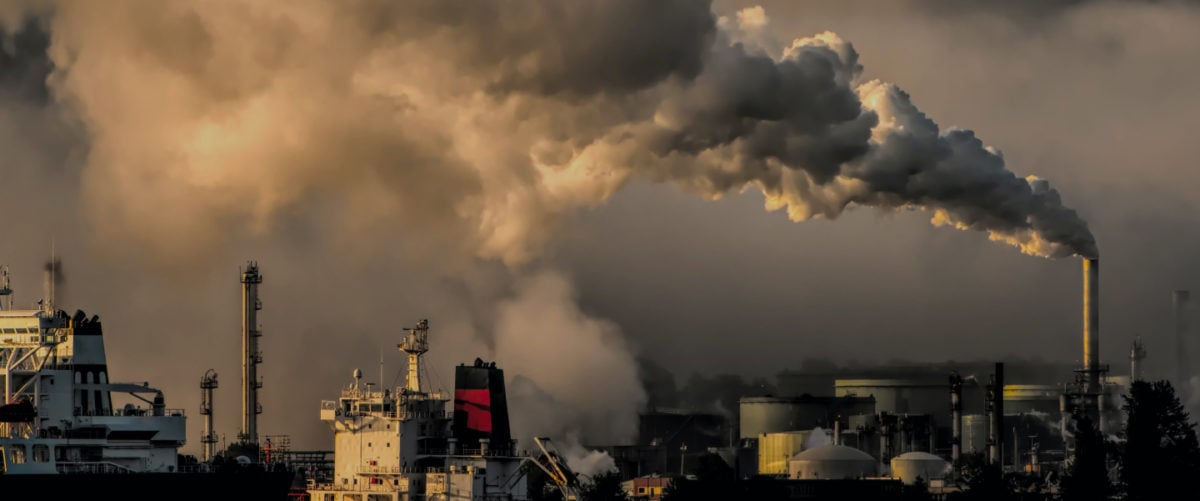 Silver smoke pours from a smokestack into gray skies and over an industrial area.