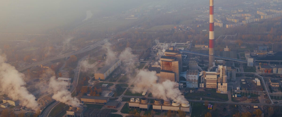 A birds eye view of a gas plant, with smoke rising from the smoke stacks and a hazy sky.