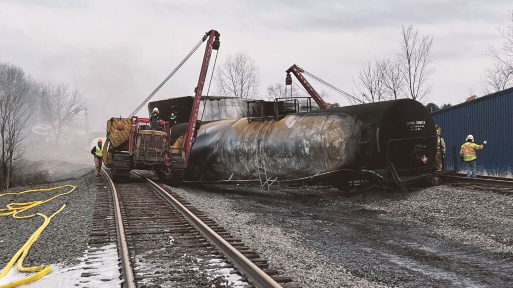 Two cranes are attached to a blackened train car sitting beside train tracks.