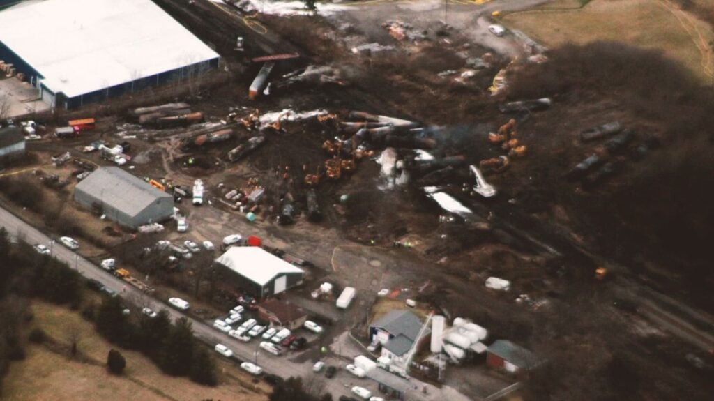 An aerial shot of the derailed train cars after the initial crash. They are lying haphazardly on blackened soil.