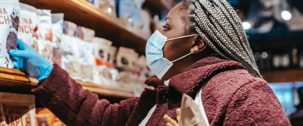 A young Black woman with braids and a mask reaches for a packet on a shelf.