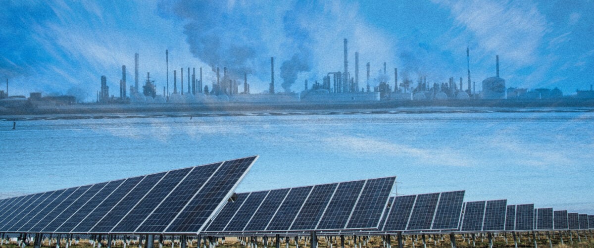 A line of smoking smoke stacks stands on the far side of a shore. A line of solar panels sits on the near side.
