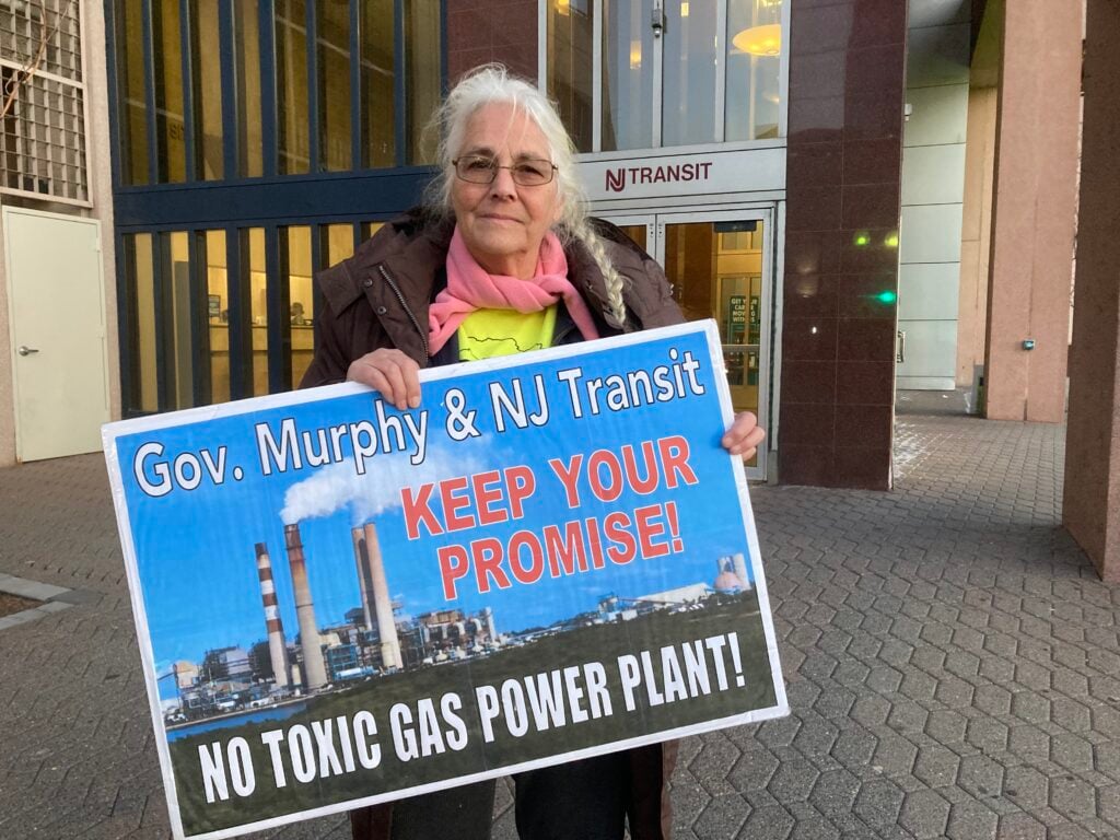 Paula Rogovin stands in front of a door marked "NJ Transit." She holds a sign that says "Gov. Murphy & NJ Transit KEEP YOUR PROMISE! NO TOXIC GAS POWER PLANT!"
