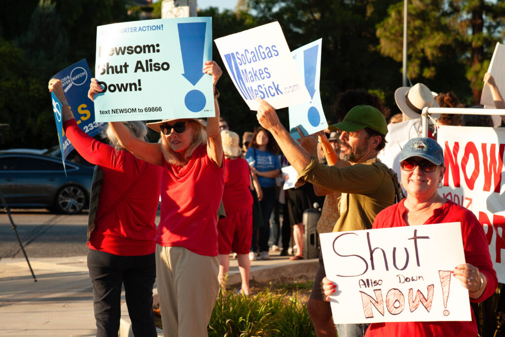 A group of people stand together holding signs that read "Newsom: Shut Aliso down!" "SoCalGas Makes Me Sick," and "Shut Aliso Down Now!"