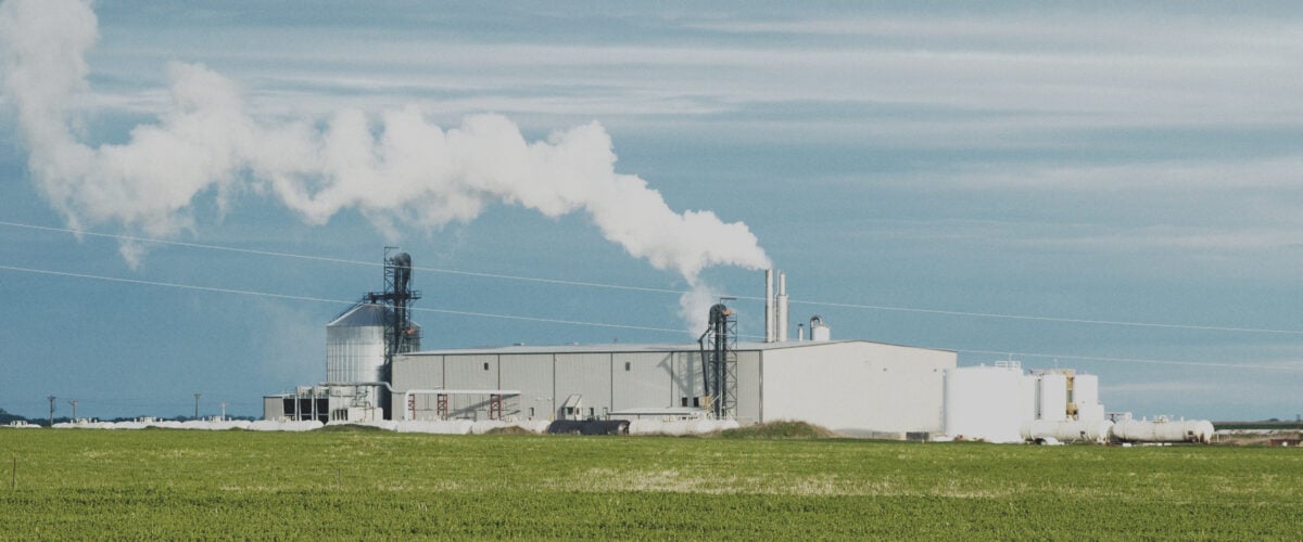 A white industrial building sits behind a green field. Smokestacks chug white clouds into a blue sky.