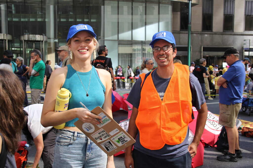 Two people wearing Food & Water Watch hats stand on the sidewalk and smile at the camera.