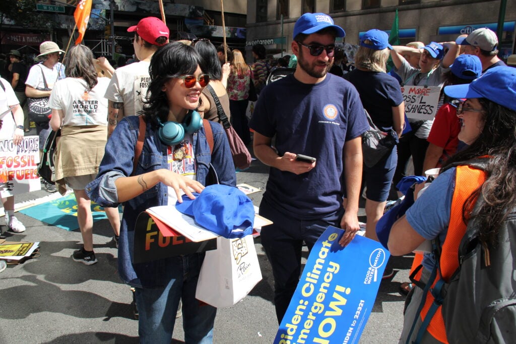 Marchers hold Food & Water Watch hats and signs while they speak to an organizer wearing a Food & Water Watch hat and shirt.