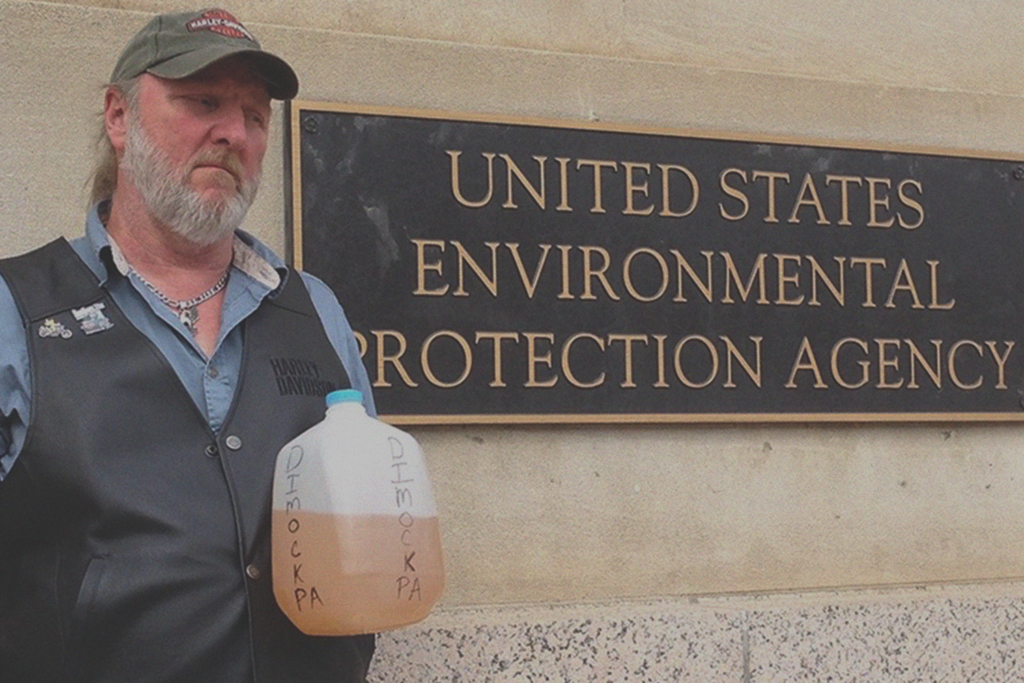 Ray Kemble, wearing a black vest and green hat, stands in front of a plaque reading "UNITED STATES ENVIRONMENTAL PROTECTION AGENCY." He holds a gallon jug of tan water labeled "DIMOCK, PA" in black letters.