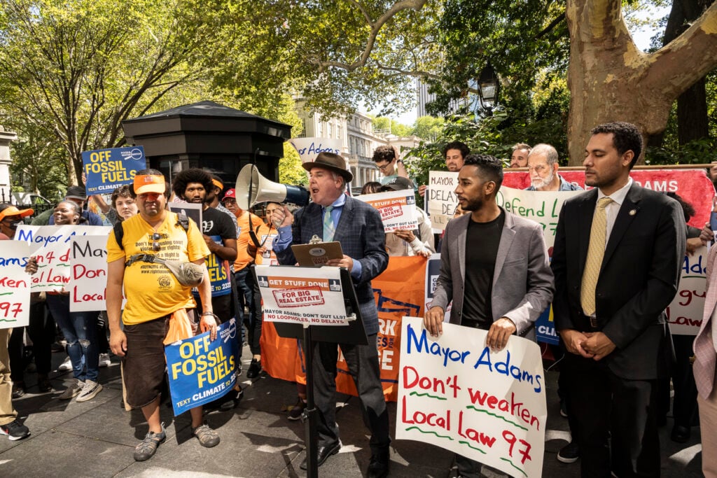 Protestors gather under trees holding signs that read "Mayor Adams Don't Weaken Local Law 97" and "Off Fossil Fuels." In the center of the group, a man behind a podium shouts into a bullhorn.