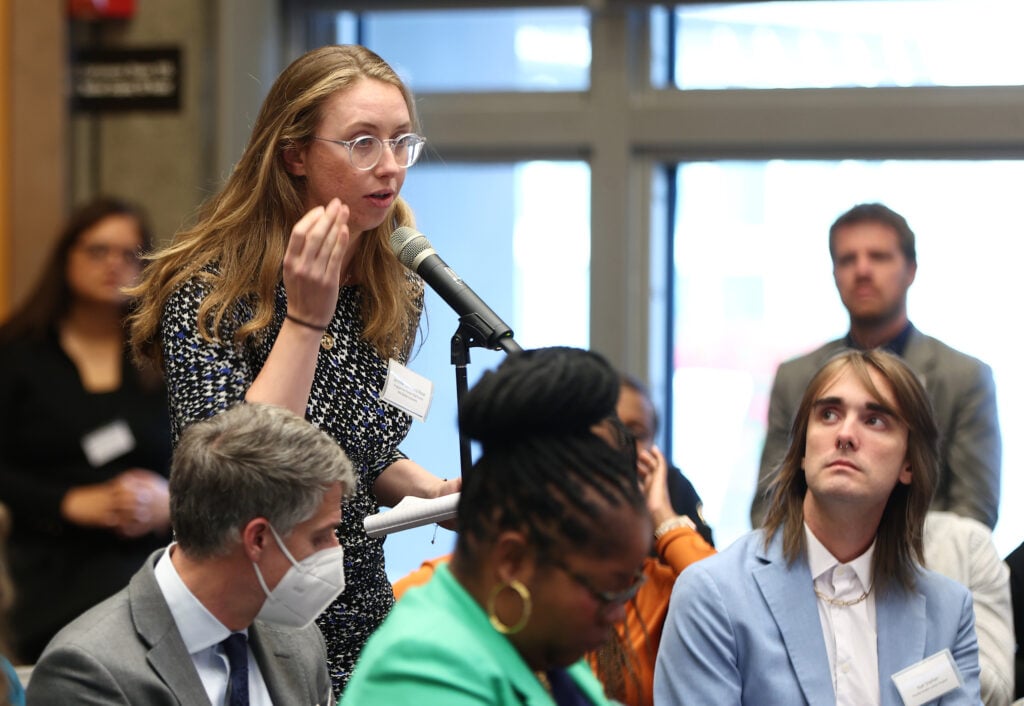 Jennifer Loveland-Rose stands and speaks in front of a microphone amongst seated people in suits.