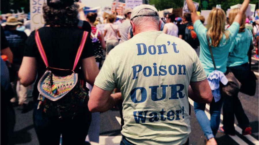 PFAS & The Chemistry of Concealment [A color photograph of a man's back at a climate march, his t-shirt reads "Don't Poison OUR Water!"]