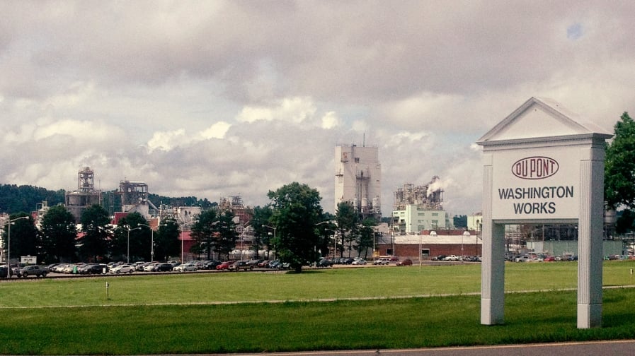 How Do PFAS Add to Environmental Racism? [a color photo depicts a grassy field in front of a full parking lot, with industrial infrastructure in the background. A sign in the foreground reads "Dupont. Washington Water Works."]