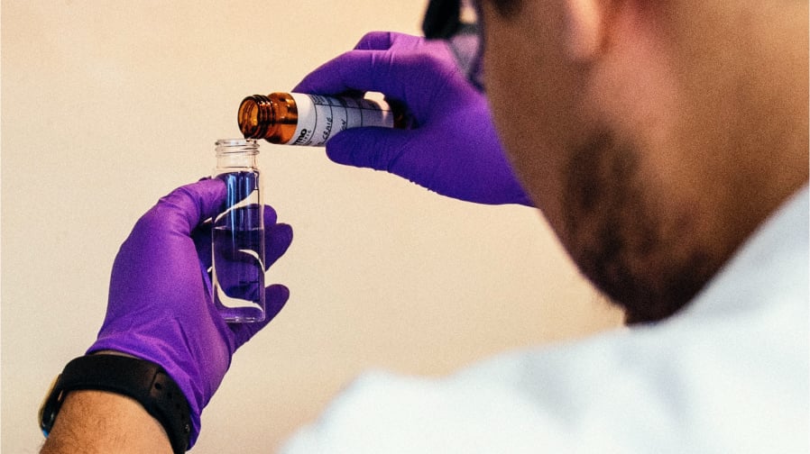 Current Federal PFAS Regulation [chemist in white lab coat can be seen from over their shoulder in this color photo. They wear purple latex gloves as they add drops of something into water to test it. 