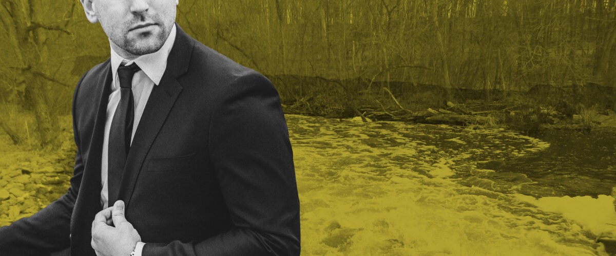A man in a suit stands in front of a stream polluted with PFAS.