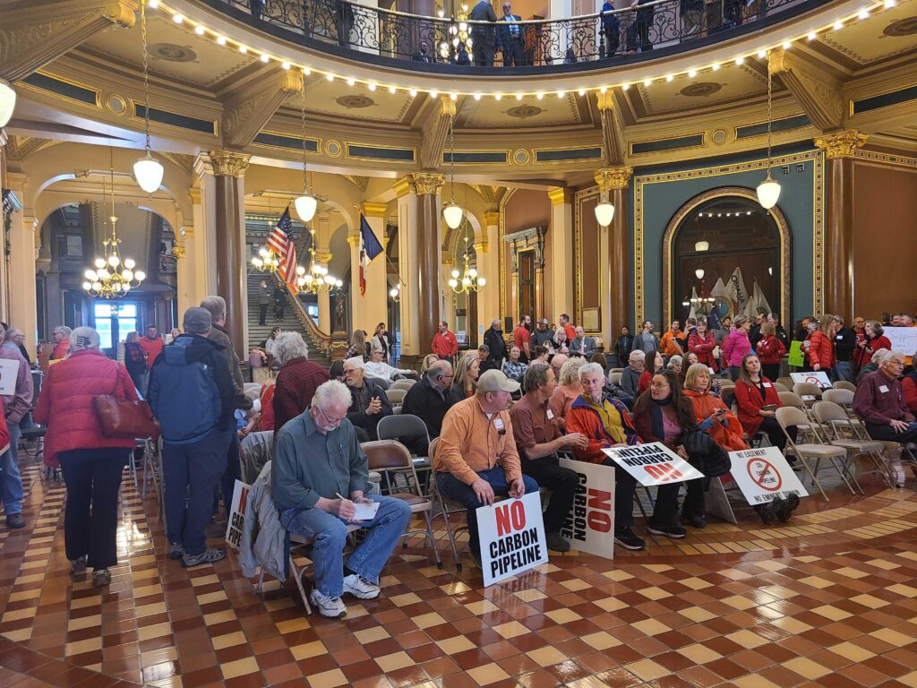 People sit in folding chairs in a large chamber with a high ceiling and gold walls. Some hold signs that read "NO CARBON PIPELINE."