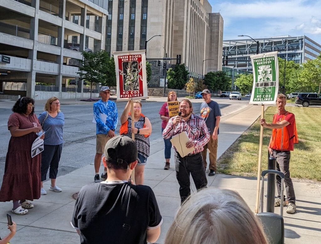 People gather on a sidewalk in Des Moines, some holding signs that say "These roots run deep. No CO2 pipelines." John Aspray speaks in the center holding a microphone.