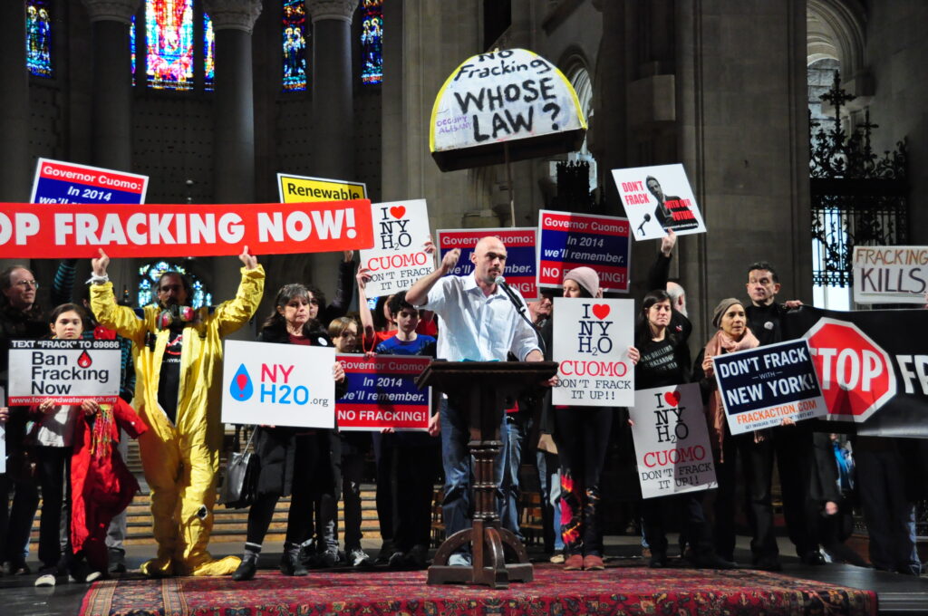 David speaks in front of a dark wooden podium. Behind him is a group of people holding protest signs that read "Ban fracking now" and "I heart NY H2O. Cuomo Don't Frack it up!" Behind them are stained glass windows and stone columns.