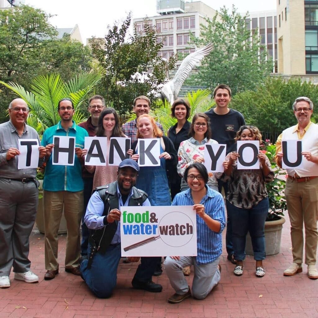 A group of staffers stand in front of green trees and plants holding signs that read "THANK YOU" and "FOOD & WATER WATCH"