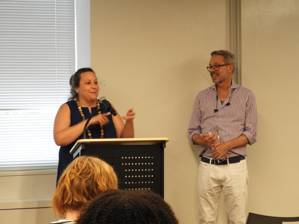 Lane stands off to the side, smiling and holding a glass award, as Patty speaks behind a podium.