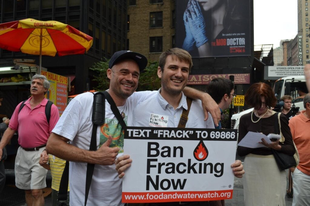 David in a t-shirt stands with his arm around Alex Beauchamp's shoulder, holding a sign that reads "Ban Fracking Now."