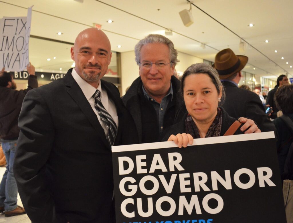 David in a black suit stands with two other people, holding a sign that reads "Dear Governor Cuomo."