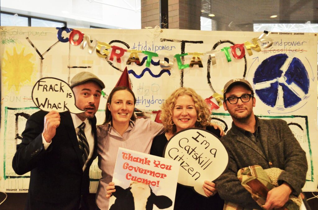 David stands with three other people under garland that spells "Congratulations." He holds a sign in the shape of a speech bubble that reads "Frack is whack."