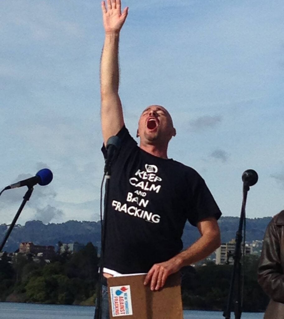 David reaches for the sky, shouting, wearing a black shirt that reads "Keep calm and ban fracking."
