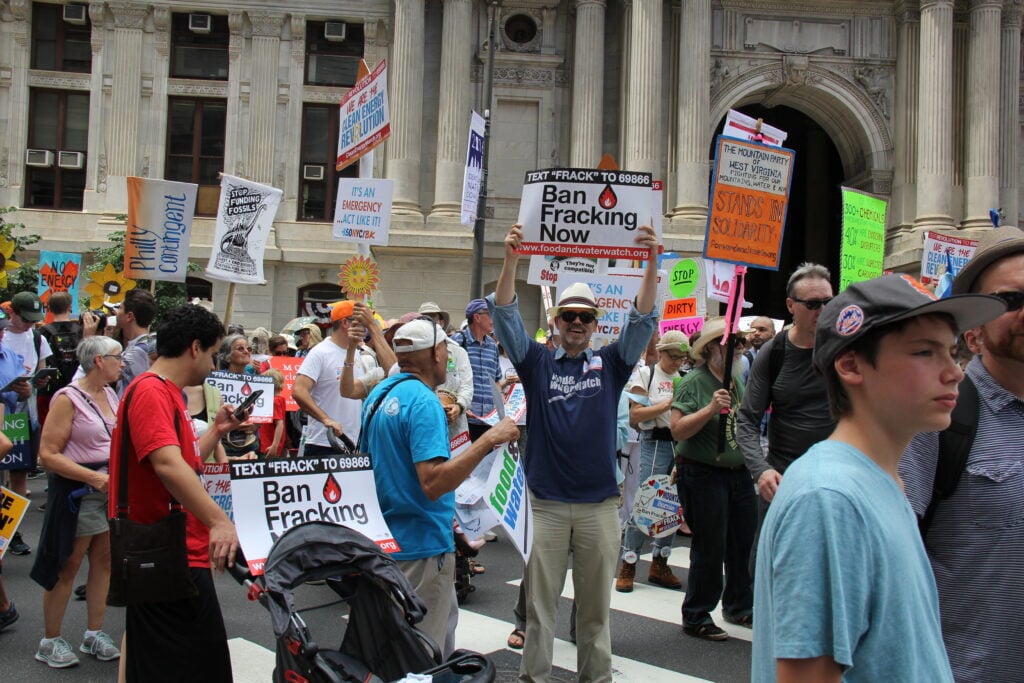 Lane stands amid the marching crowd, smiling and holding up a sign that reads "Ban Fracking Now."