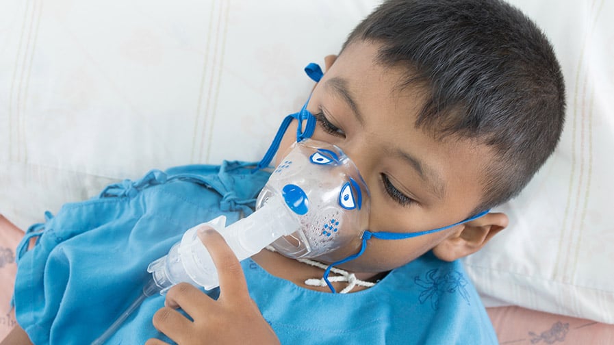 A toddler lays in bed with a nebulizer mask on their face, which is delivering medicine for their asthma symptoms.