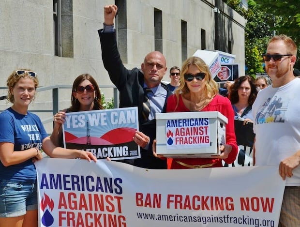 David wears a suit and has his arm raised, hand clenched in a fist. He stands with four other people holding signs that read "Americans Against Fracking."