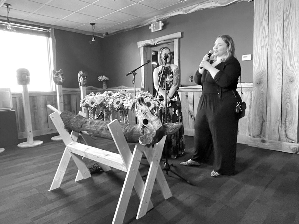 Hilary stands to the right speaking into a microphone, in front of a statue of a deer head carved from wood.