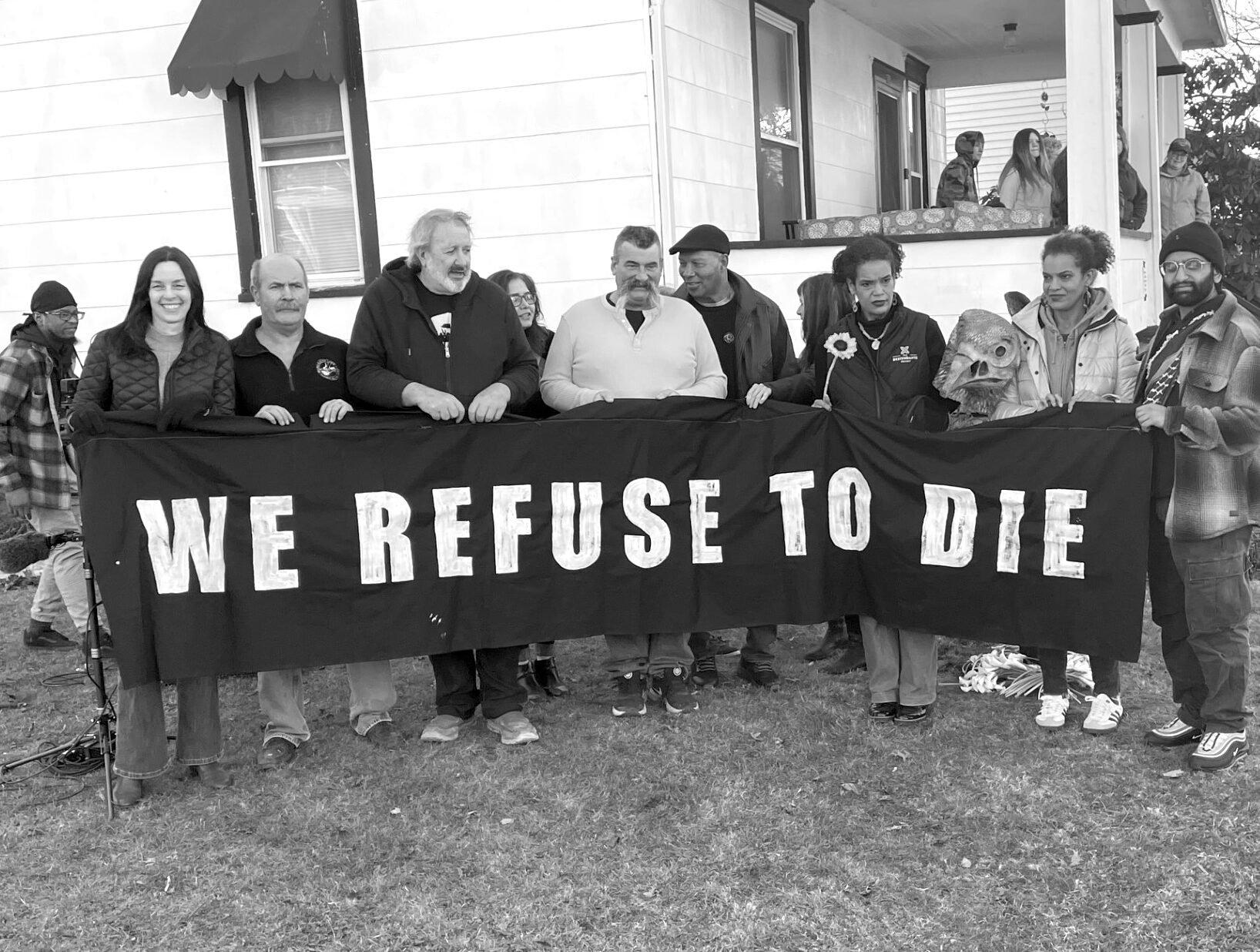 Seven people gather on a lawn on the side of a white house, holding a black banner that reads "WE REFUSE TO DIE."