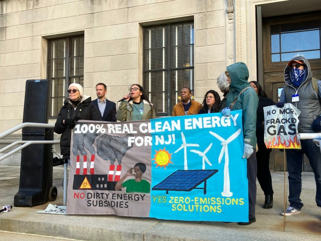 Seven speakers stand on stone steps outside of the New Jersey Capitol Building, holding a banner reading: "100% REAL CLEAN ENERGY FOR NJ. NO DIRTY ENERGY SUBSIDIES. YES ZERO-EMISSIONS SOLUTIONS."