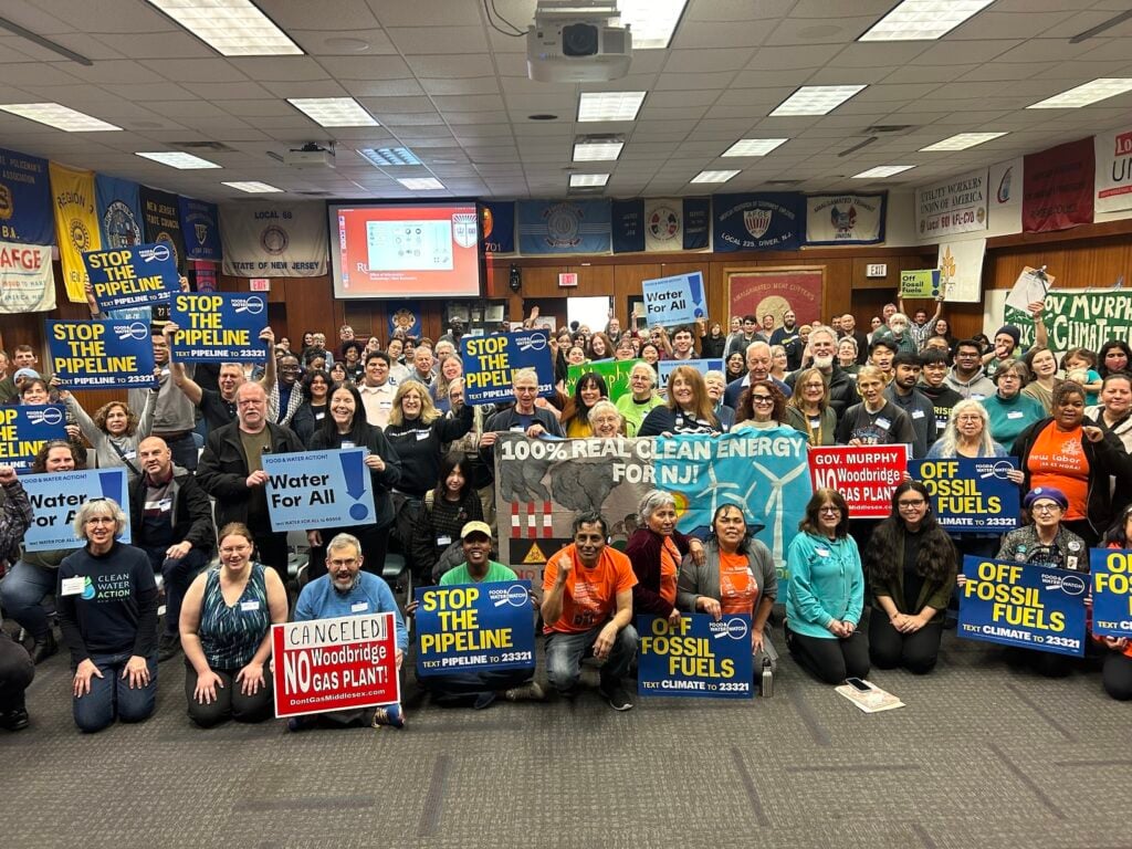 Dozens of activists gather in a large room with brown walls and grey carpet, holding signs that way "Stop the pipeline," "100% real clean energy for NJ!" "Off fossil fuels," and more.