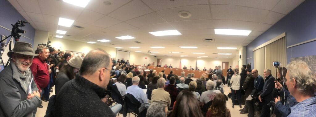 A few inside the Franklin Park Borough Council building from the back. People sit in rows facing a high table where the Council members sit. People stand along the sides.