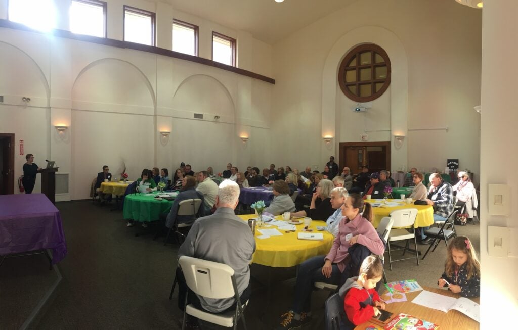 Dozens of residents gather around circular tables in a large white room with high ceilings.