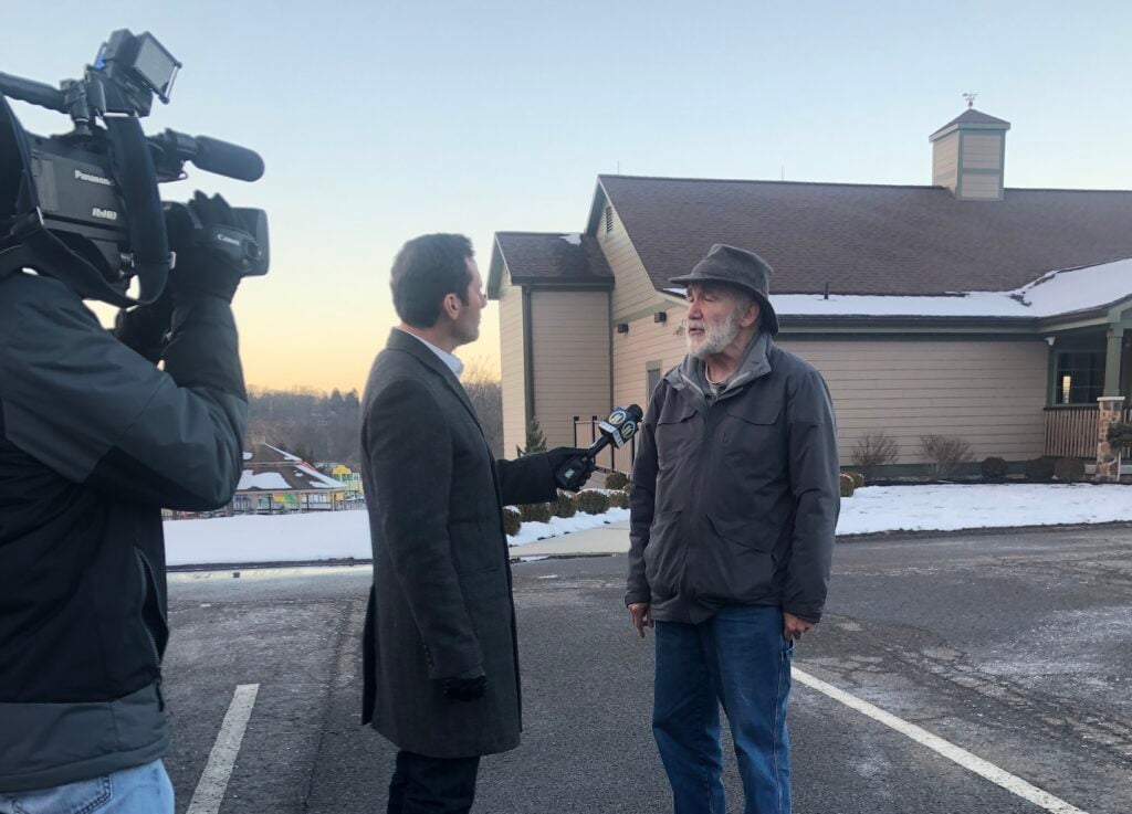In a parking lot in front of a small building lightly dusted with snow, Thaddeus speaks to a reporter holding a mic. A camera person points a large black camera in their direction.