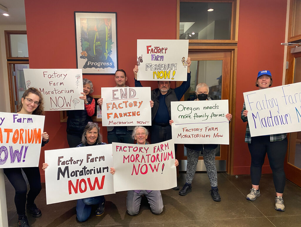 Eight people stand in front of a red wall holding signs that read "Factory farm moratorium now!" and "End factory farming."