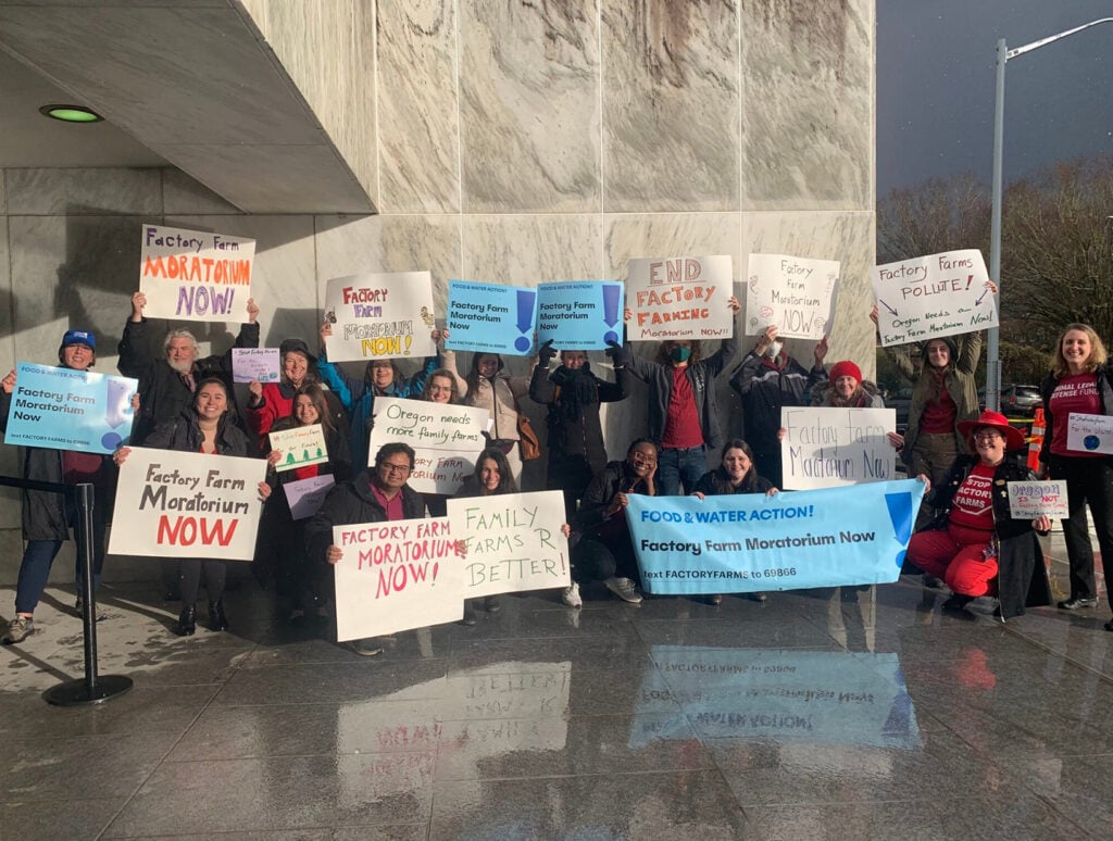 A group of people stand in front of a stone wall outdoors holding signs that read "Factory Farm Moratorium now."