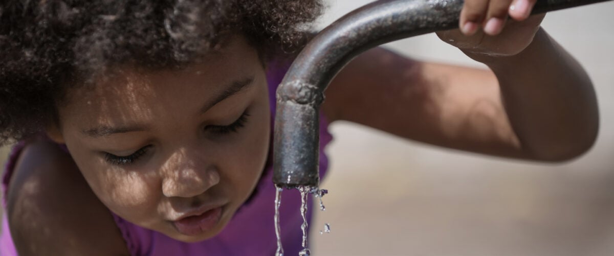 A young Black girl leans toward a running faucet for a drink.
