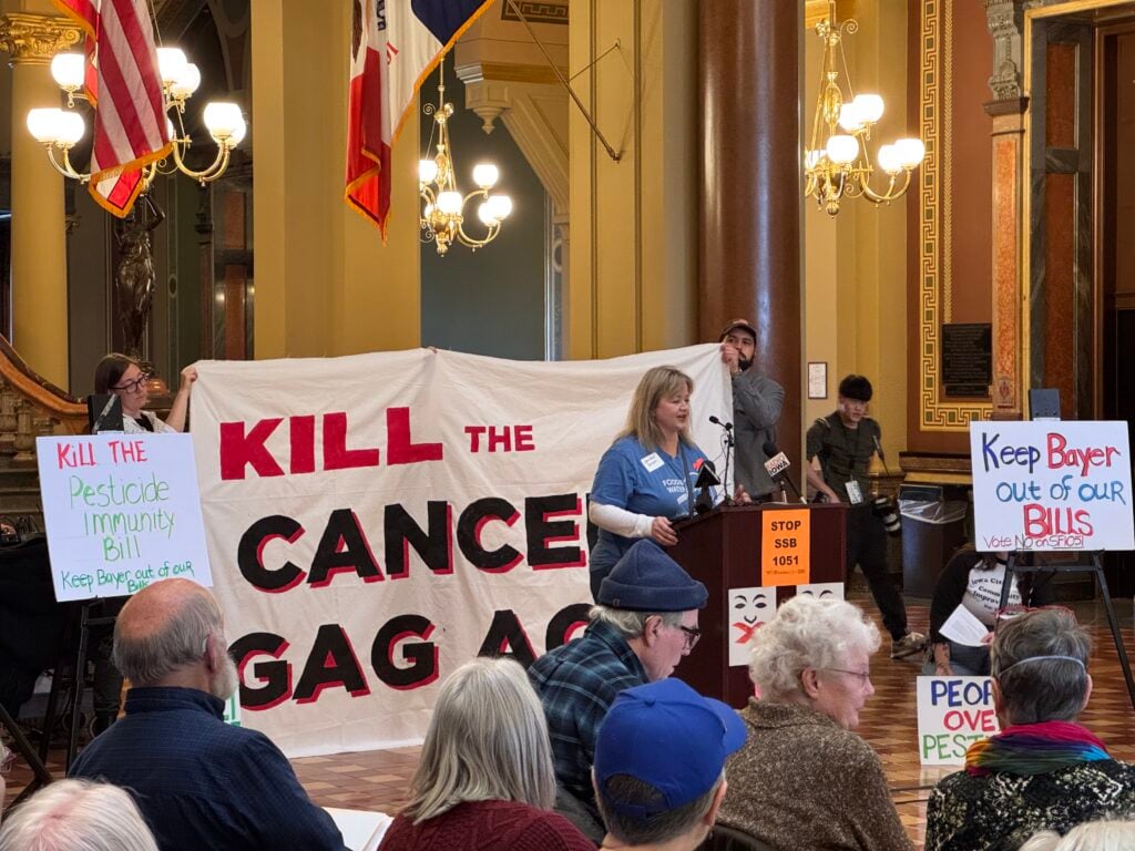 A woman wearing a Food & Water Watch t-shirt addresses a crowd in the state capitol rotunda with a banner behind her reading "Kill the Cancer Gag Act."
